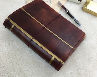 May include: A brown leather journal with gold accents and a burgundy elastic closure. The journal is closed and lying on a white surface.