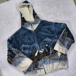 May include: A zip-up hooded jacket made of soft blue and white fleece. The fabric features a winter landscape print with snow-covered cabins, trees, and mountains. The interior of the hood and the sleeve cuffs are lined with a solid cream-colored fleece.