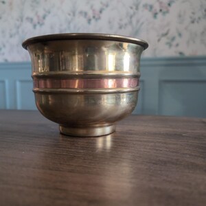 May include: A polished brass bowl featuring a distinct horizontal copper band encircling the middle section. The bowl has a wide, flared rim, a smooth metallic finish, and a circular base, presenting a two-toned appearance with gold and copper hues.