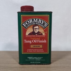 Puede incluir: Una lata metálica rectangular de acabado de aceite de tung tradicional Formby's Furniture Workshop con una tapa de rosca roja oscura. La etiqueta es verde y roja, con una ilustración de una persona con gafas y el texto 473 mL.
