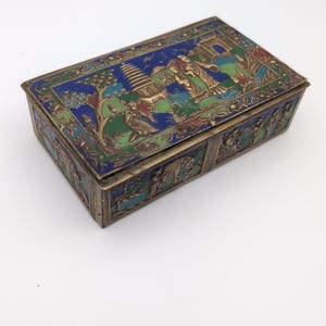 May include: A rectangular metal box featuring intricate cloisonné enamel work in shades of blue and green. The lid and sides are decorated with embossed scenes depicting a pagoda, trees, and figures. The metal has a tarnished brass-toned finish.