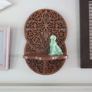 Peut inclure: Étagère murale en bois avec un motif sculpté complexe. Une petite figurine de chien vert clair est posée sur l'étagère. L'étagère est fixée à un mur blanc, avec un cadre rose et un cadre foncé visibles.