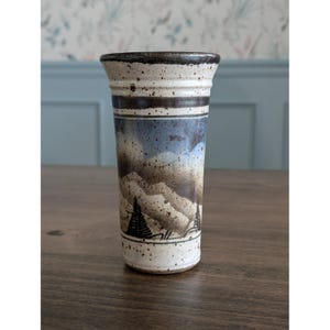 Pode incluir: Um vaso de cerâmica alto e cilíndrico com um design de paisagem. O vaso apresenta uma base creme salpicada, uma faixa marrom escuro e uma cena pintada de montanhas em tons de azul e marrom. A borda é marrom escuro.