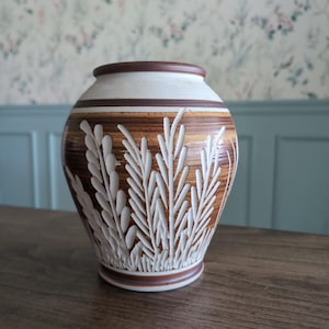 Puede incluir: Un jarrón decorativo de cerámica con un esquema de color marrón, blanco y beige. El jarrón presenta un diseño texturizado de hojas blancas y un borde marrón. El jarrón mide aproximadamente 20 cm de alto.