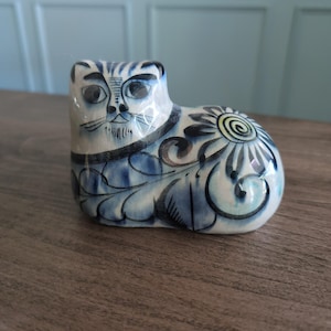 May include: A ceramic cat ornament with a blue and white floral pattern. The cat has large eyes and a detailed face. The figurine is approximately 10 cm long and 8 cm tall.