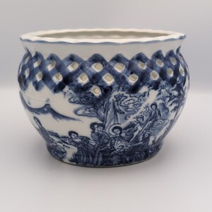 May include: A white ceramic bowl featuring a blue hand-painted landscape scene with figures and trees. The upper portion of the bowl has a decorative lattice cut-out pattern, while the rim is scalloped. The base is rounded and tapers inward.