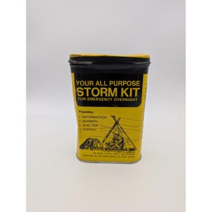 Puede incluir: Una lata metálica rectangular amarilla y negra etiquetada como YOUR ALL PURPOSE STORM KIT FOR EMERGENCY OVERNIGHT. El frente presenta un gráfico de una persona sentada junto a una fogata y una tienda, con texto que enumera información, calor, refugio, energía.