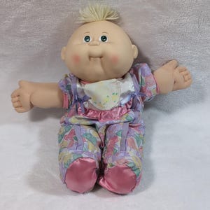 Puede incluir: Una muñeca vintage de Cabbage Patch Kids con cabeza calva y mechón rubio. La muñeca viste un conjunto de estampado floral pastel con detalles en rosa y un babero. Los ojos de la muñeca son verdes y sus mejillas son rosadas.