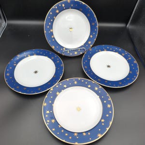 May include: A set of four round ceramic plates featuring a white center with a single gold star and a wide dark blue rim decorated with numerous gold stars of varying sizes. Each plate has a thin gold-colored trim along the outer edge.