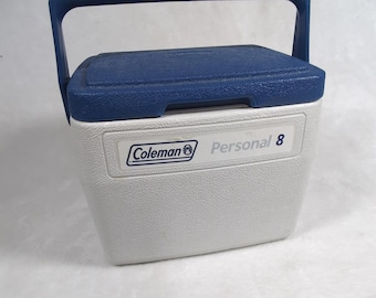 Puede incluir: Una nevera portátil Coleman Personal 8 de color blanco y azul con exterior de plástico texturizado, tapa abatible y un asa de transporte azul resistente. El frente presenta una etiqueta rectangular con el logotipo de Coleman y el texto Personal 8 en azul.