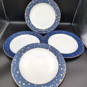 May include: A set of four round ceramic plates featuring a solid white center and a wide dark blue rim. The blue border is decorated with various gold-colored star shapes of different sizes and styles, finished with a thin gold edge.