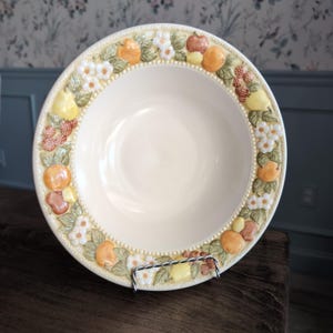 May include: A round ceramic bowl with a cream-colored center and a wide, raised rim featuring a relief pattern of colorful fruits, including oranges, pears, and berries, interspersed with white blossoms and green leaves on a textured border.