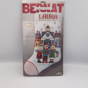 May include: A Bernat Counted CrossStitch kit for a Christmas stocking featuring the name LAURA. The design shows three carolers in winter clothing standing in front of a village building with blue, red, and green details under falling snowflakes.