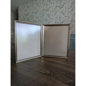 May include: A gold-colored, hinged picture frame with a decorative border. The frame is open, revealing two blank spaces for photos. The frame is sitting on a dark wooden surface.