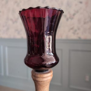 May include: A deep burgundy glass candle holder with a flared, scalloped rim and a smooth, curved body. The glass piece is securely mounted atop a light-toned, turned wooden base that features a series of horizontal ridges and a tapered neck.