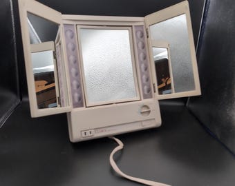 May include: A beige tri-fold vanity mirror with integrated lighting panels on the sides. The central mirror is flanked by two adjustable side panels. A control dial and a power switch are located on the base, with the brand name Clarice visible.