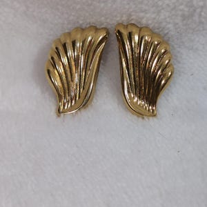 May include: A pair of gold-coloured earrings. Each earring features a fan-like design with vertical ridges. The earrings are presented on a white, textured surface. The earrings are likely made of metal and are designed to be worn on the ear.