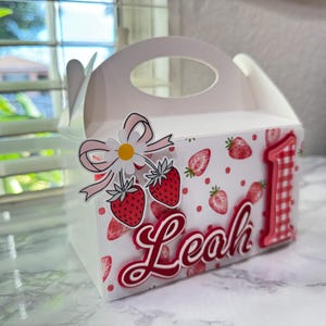Puede incluir: Una caja de regalo blanca con asa, decorada con fresas, una margarita y el nombre "Leah" en escritura roja. Tambi&eacute;n aparece el n&uacute;mero rojo "1", lo que sugiere un tema de primer cumplea&ntilde;os.