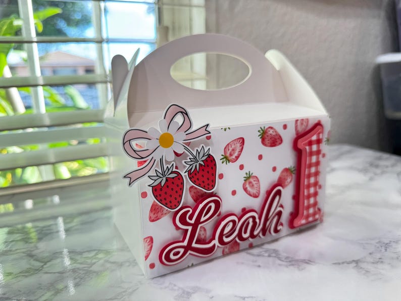 Puede incluir: Una caja de regalo blanca con asa, decorada con fresas, una margarita y el nombre "Leah" en escritura roja. Tambi&eacute;n aparece el n&uacute;mero rojo "1", lo que sugiere un tema de primer cumplea&ntilde;os.