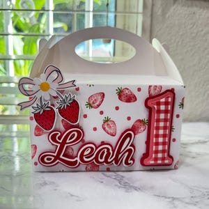Puede incluir: Una caja de regalo blanca con asa, decorada con fresas rojas y el nombre "Leah". El n&uacute;mero rojo "1" con estampado de cuadros rojos y blancos est&aacute; en el lateral, con una decoraci&oacute;n de margarita y fresa.