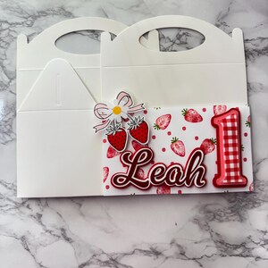 Puede incluir: Caja blanca para recuerdos de fiesta con asa, decorada con un tema de fresas. La caja presenta una tarjeta con fresas, el nombre "Leah" y el n&uacute;mero "1" en rojo. Un lazo y flores acent&uacute;an el dise&ntilde;o.