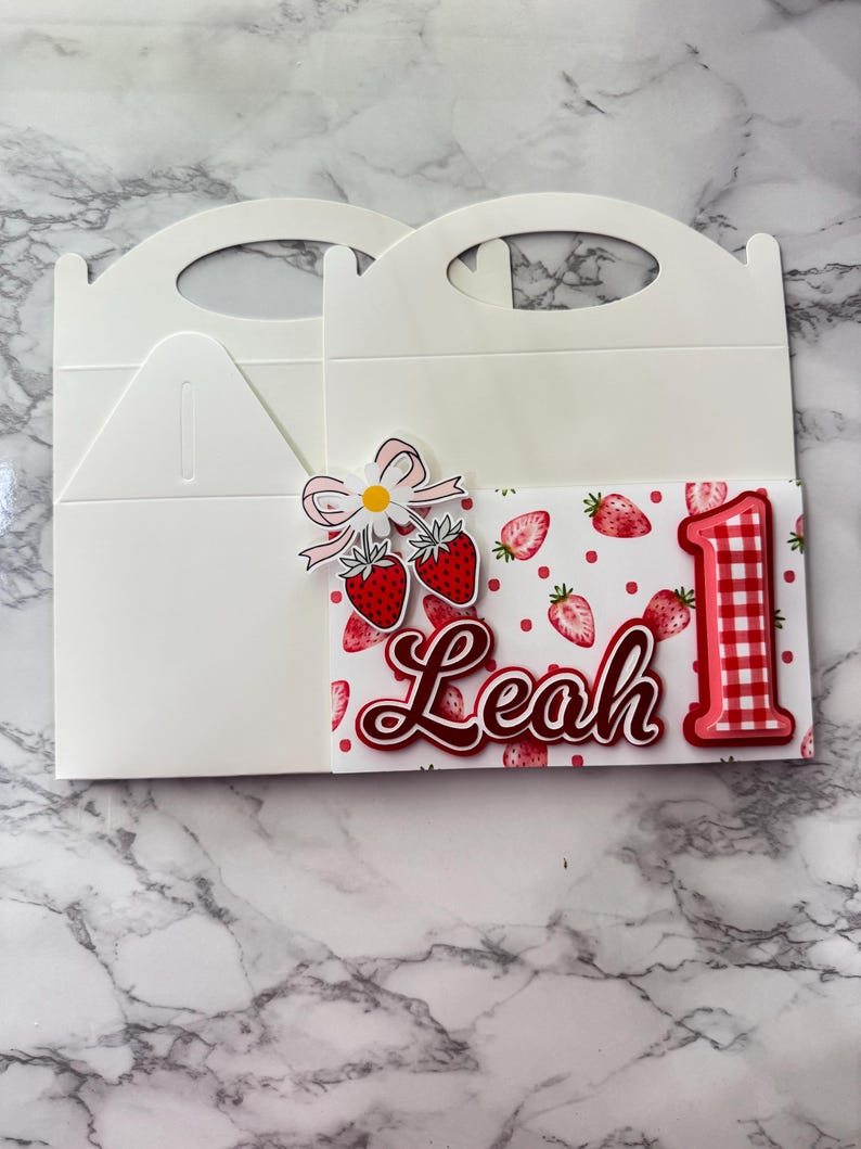 Puede incluir: Caja blanca para recuerdos de fiesta con asa, decorada con un tema de fresas. La caja presenta una tarjeta con fresas, el nombre "Leah" y el n&uacute;mero "1" en rojo. Un lazo y flores acent&uacute;an el dise&ntilde;o.