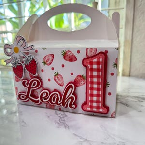 Puede incluir: Una caja de regalo rosa claro decorada con fresas, lunares y el nombre "Leah" en escritura roja. El n&uacute;mero "1" rojo grande con un patr&oacute;n de cuadros est&aacute; en la parte delantera, junto con un adorno de fresa y margarita.