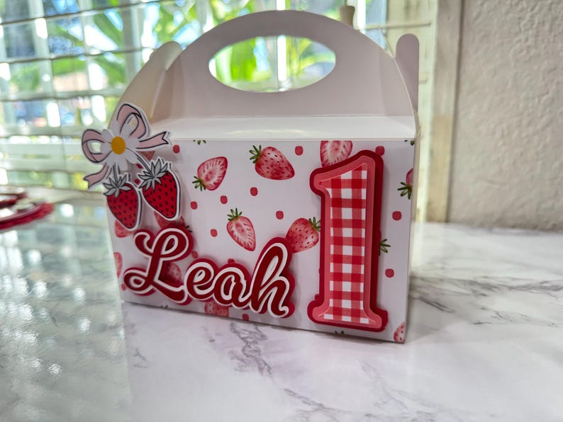 Puede incluir: Una caja de regalo rosa claro decorada con fresas, lunares y el nombre "Leah" en escritura roja. El n&uacute;mero "1" rojo grande con un patr&oacute;n de cuadros est&aacute; en la parte delantera, junto con un adorno de fresa y margarita.