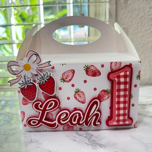 Puede incluir: Caja blanca con forma de caba&ntilde;a decorada con fresas rojas y el nombre "Leah" en cursiva. Un gran n&uacute;mero "1" en cuadros rojos y blancos y un adorno de fresa y margarita tambi&eacute;n est&aacute;n presentes.