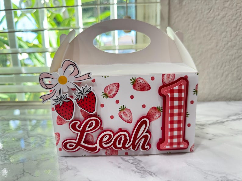 Puede incluir: Caja blanca con forma de caba&ntilde;a decorada con fresas rojas y el nombre "Leah" en cursiva. Un gran n&uacute;mero "1" en cuadros rojos y blancos y un adorno de fresa y margarita tambi&eacute;n est&aacute;n presentes.
