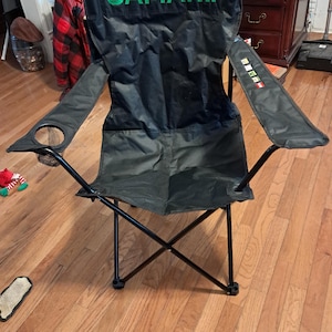 May include: Black folding camping chair with the name "JAMARII" in green lettering on the backrest. The chair has a built-in cup holder and armrests. The frame is black, and the chair is set up on a wooden floor.