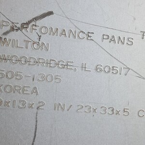 May include: Close-up of a metal baking pan with the text "PERFORMANCE PANS" and "WILTON". The pan's dimensions are listed as 9x13x2 inches, equivalent to 23x33x5 cm.