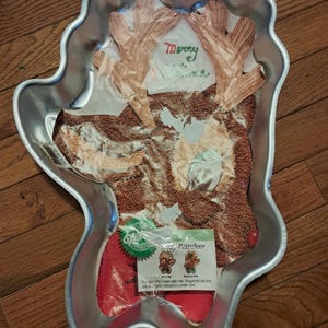 May include: A silver-colored, reindeer-shaped cake pan with a decorative insert. The insert features a reindeer face with brown fur and antlers, and the word "Merry" in green. A red cake liner and instructions are also included.