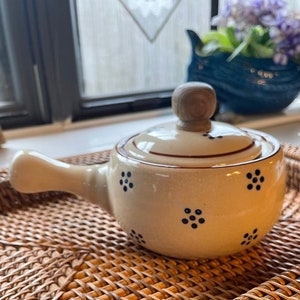 May include: A white ceramic teapot with a brown lid and a wooden knob. The teapot has a handle and is decorated with blue and white polka dots. The teapot is sitting on a woven wicker tray.
