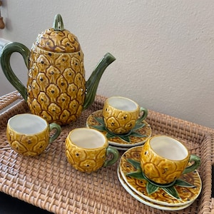 May include: A pineapple-shaped teapot and four matching teacups and saucers. The teapot and cups are decorated with a realistic pineapple design in shades of yellow, green, and brown. The set is displayed on a woven wicker tray.