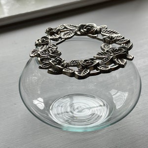 May include: A clear glass vase with a silver metal floral design lid. The lid is a wreath of roses and leaves.