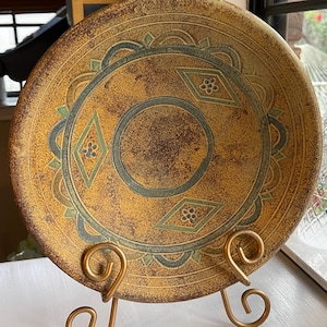 May include: A round ceramic plate with a textured mustard yellow and brown mottled finish. The plate features a central circle, three diamond shapes with small flower motifs, and a scalloped border in muted green, displayed on a gold metal stand.