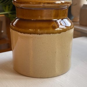 May include: A cylindrical ceramic crock featuring a two-tone design with a glossy dark brown glazed upper section and a matte light beige lower body. The vessel has a wide, slightly flared rim and a smooth, uniform exterior surface finish.