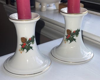 May include: Two white ceramic candle holders with gold trim, each holding a tall, pink candle. The holders feature a festive design of holly leaves, red berries, and a pinecone. A blue and white planter is in the background.