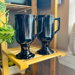 May include: Two black ceramic mugs with handles and a pedestal base. The mugs are on a wooden shelf.