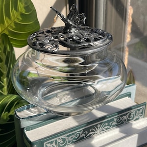 Op de afbeelding: Een heldere glazen kom met een ronde basis en een afneembaar zilverkleurig metalen deksel. Het deksel is voorzien van een gedetailleerd reliëfontwerp van een vlinder die op een cluster van bloemen en bladeren rust als afdekking voor de kom.