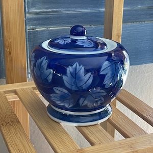 May include: A blue and white ceramic jar with a lid. The jar has a floral design and is sitting on a wooden shelf.