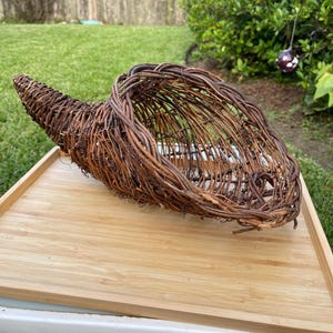 May include: A cornucopia basket constructed from dark brown, intertwined natural twigs and branches. The woven structure features an open, hollow interior with a tapered, curved tail end and a wide, flared opening at the front.