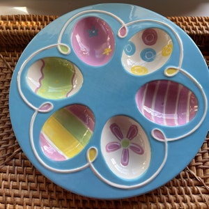 May include: A light blue ceramic Easter egg plate with six egg-shaped indentations. Each indentation is decorated with a different colorful Easter egg design.