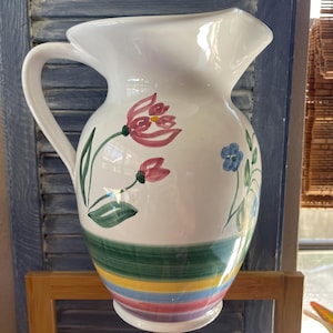 May include: White ceramic pitcher with a floral design featuring pink and blue flowers. The pitcher has a green, yellow, blue, and pink striped pattern around the bottom.