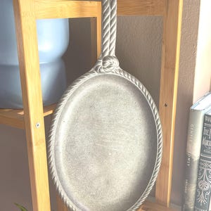 May include: Oval, silver-colored serving tray with a rope-like handle and border. The tray is made of metal and has a slightly textured surface. The handle is attached to the tray with a knot design. The tray is displayed on a wooden shelf.