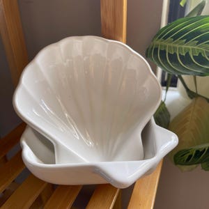 May include: A white ceramic seashell-shaped dish. The dish has a scalloped edge and a smooth, glossy finish. It is sitting on a wooden shelf. The dish is designed to hold small items or serve as a decorative piece.