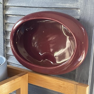 May include: A burgundy-colored, oval-shaped ceramic planter. The planter has a glossy finish and a smooth interior. It is sitting on a light brown wooden shelf, with a gray shutter background.