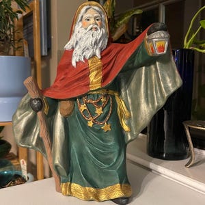 May include: A ceramic figurine of a bearded man wearing a red and green robe, holding a staff and a lantern. He has a white beard and a gold chain with stars around his neck.