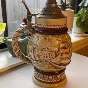 May include: A ceramic beer stein with a detailed scene of a ship and buildings. The stein has a brown, red, and white color scheme and a metal lid with a rope design around the top.